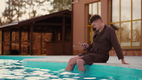 Ein-Entspannter-Mann-Ruht-Sich-Im-Freibad-Eines-Parks-Aus-Und-Surft-Mit-Einem-Modernen-Smartphone-In-Den-Sozialen-Medien